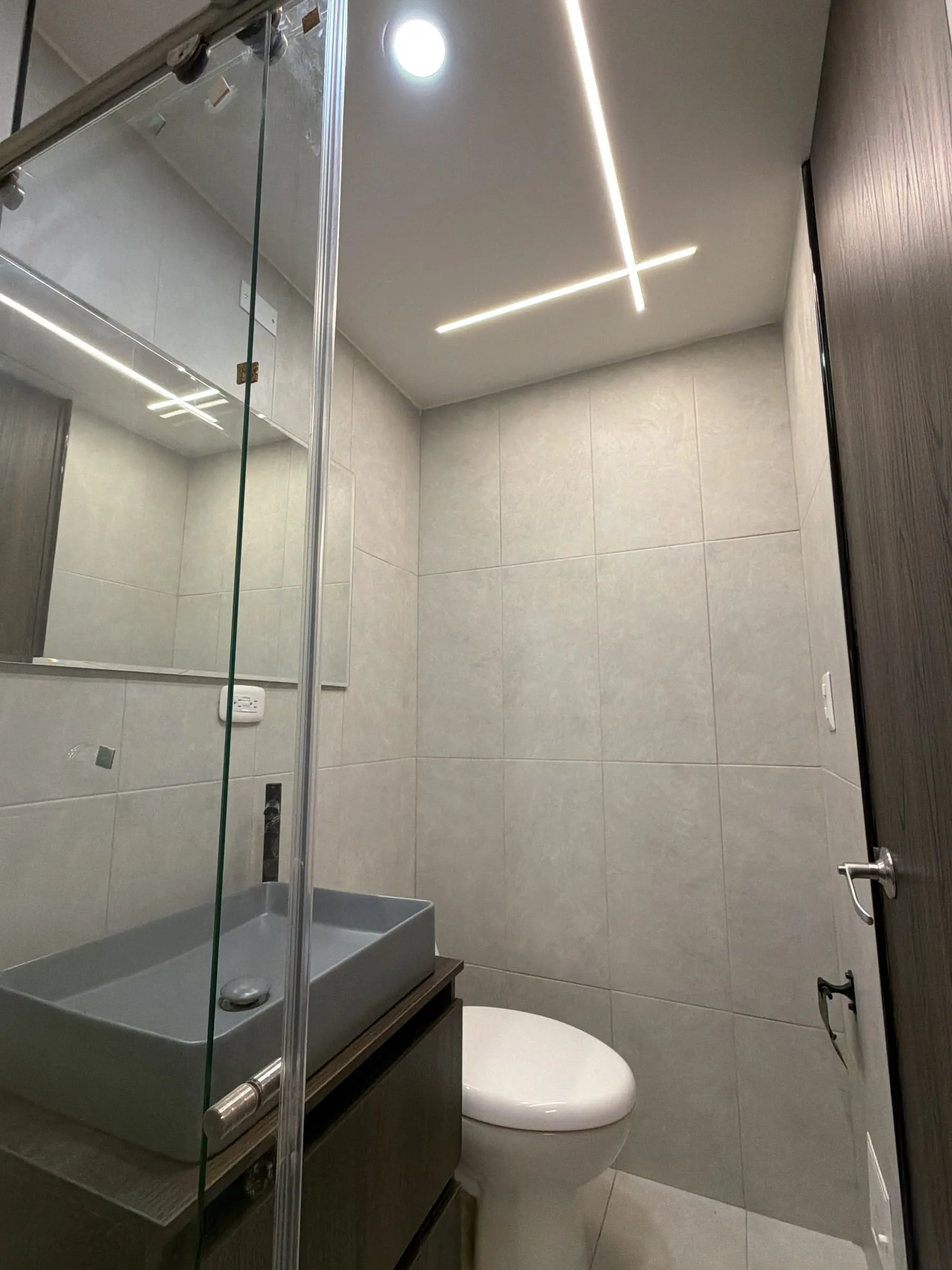 Baño con luz lineal cruzada en el cielo raso y ducha en vidrio templado.