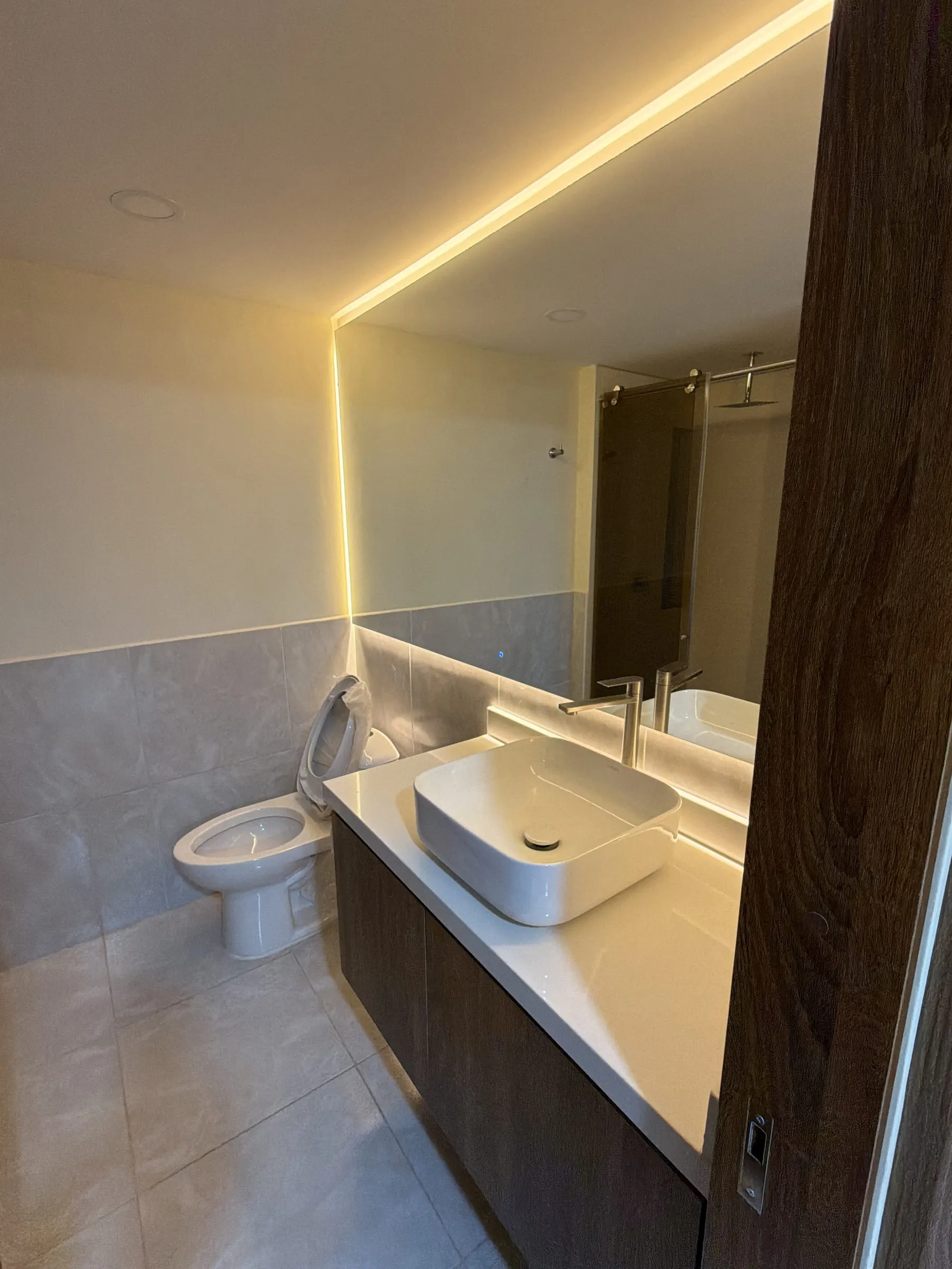 Baño con espejo retroiluminado, lavamanos sobrepuesto y mueble en madera bajo una celosía de listones.