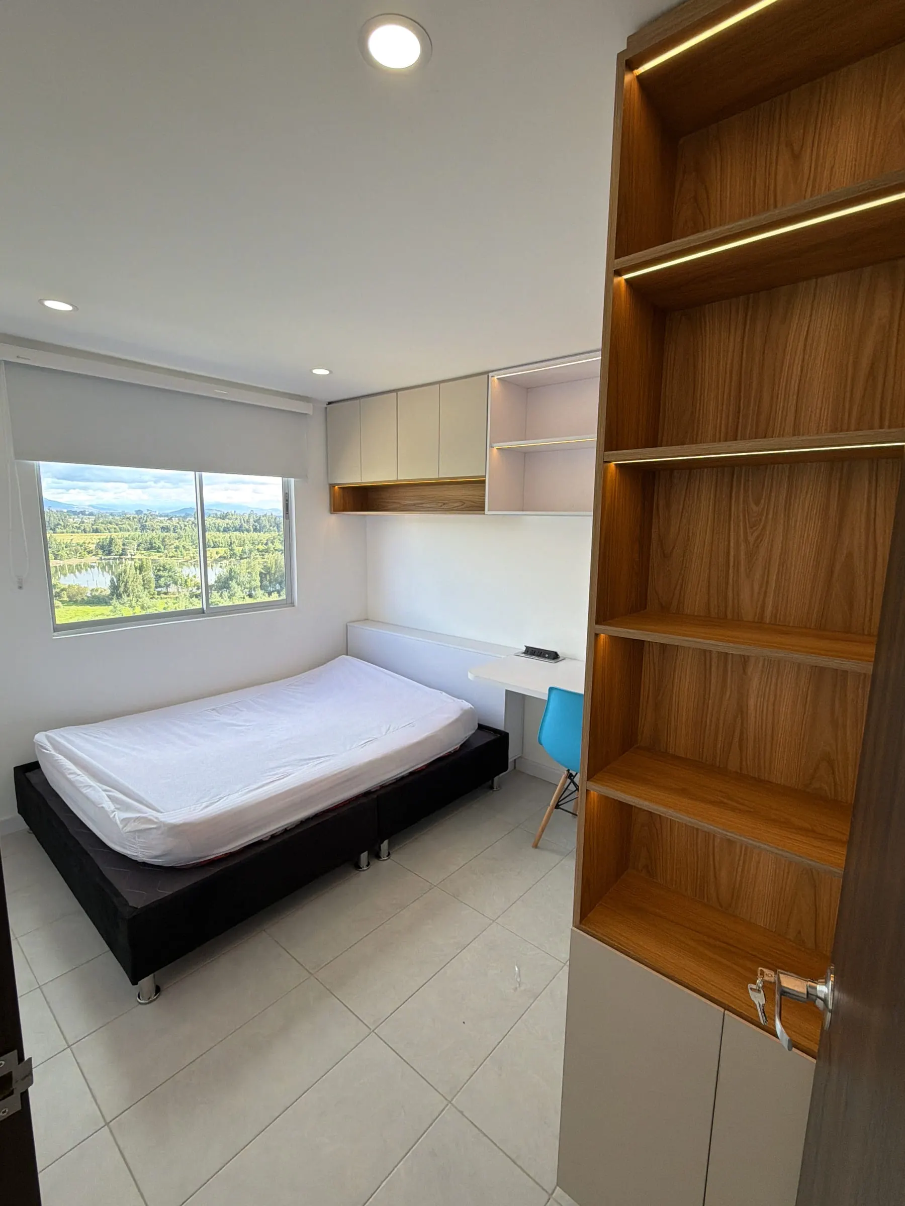 Habitación con la cama junto a la ventana y una columna de repisas en madera iluminada.
